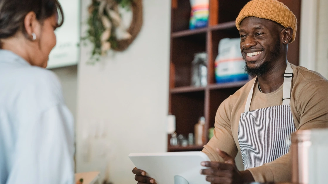 Smiling shop owner in an apron serving a customer at the counter with a tablet