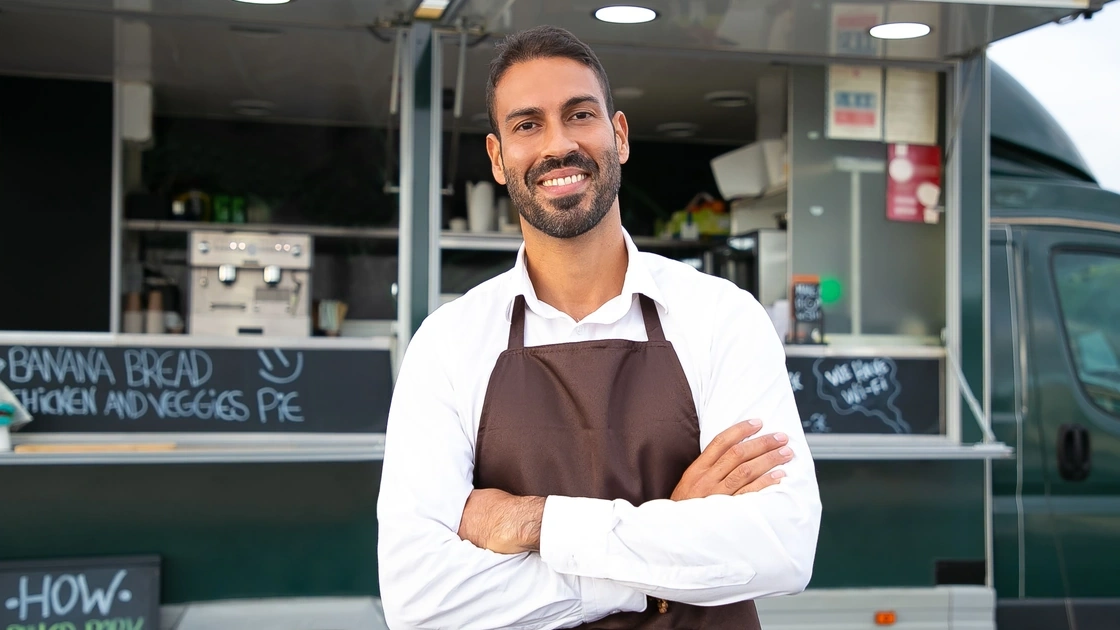 Smiling food truck owner in an apron standing with arms crossed outside his truck