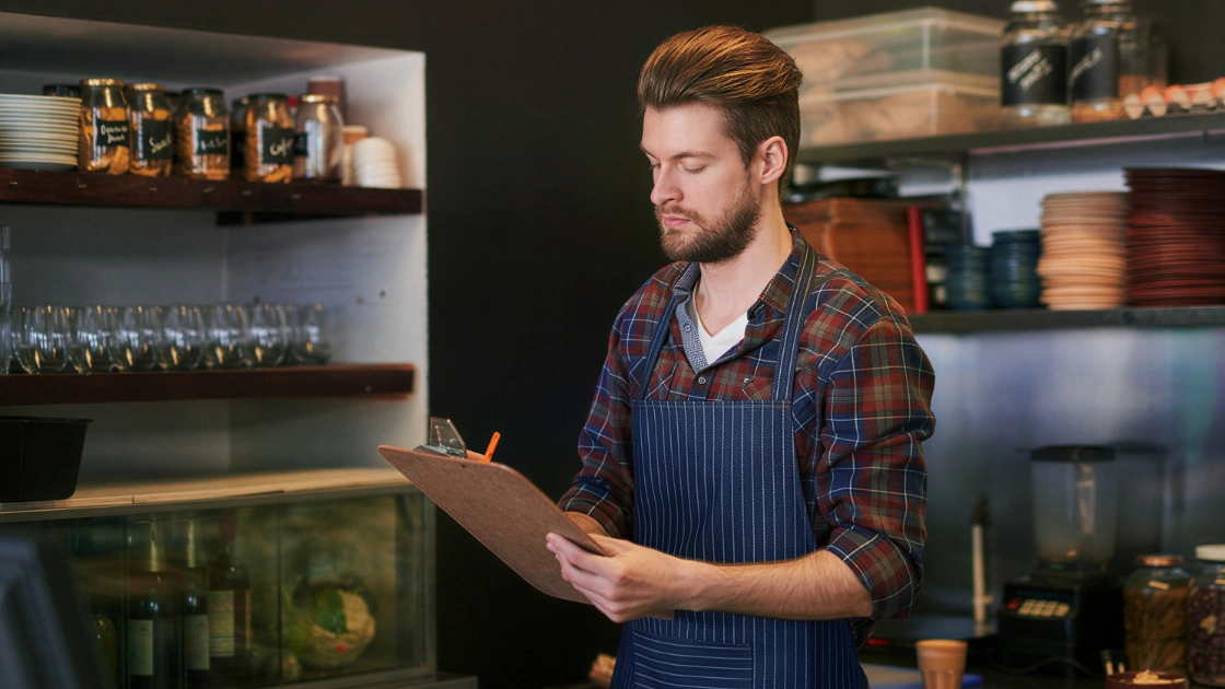 Café owner in an apron checking stock on a clipboard behind the counter