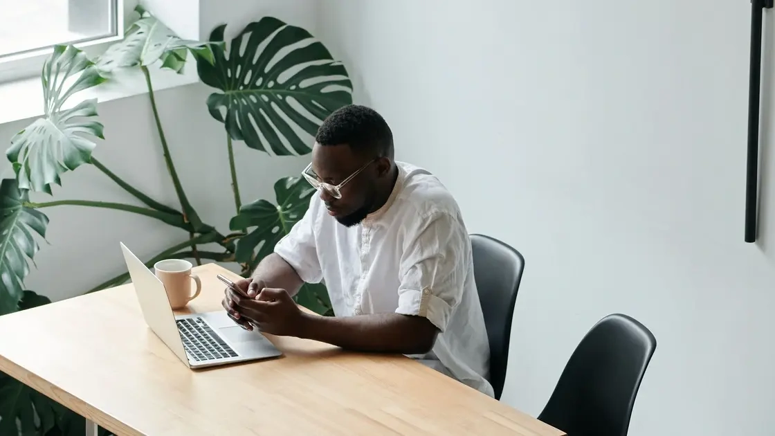 Man using his phone at a wooden desk with a laptop and a monstera plant